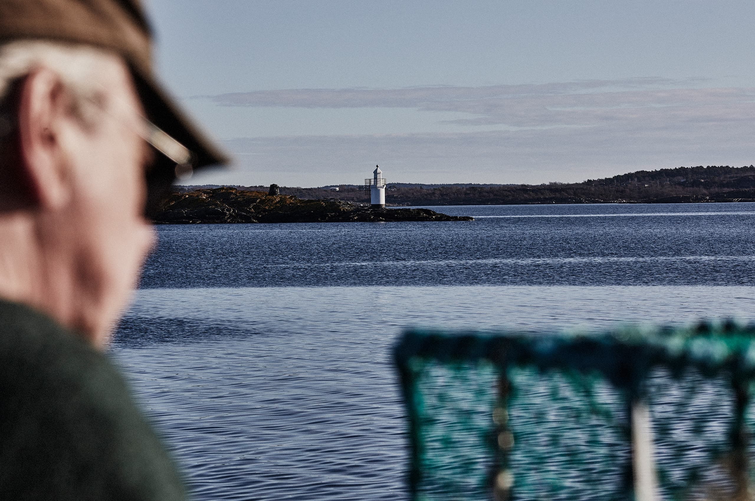 äldre man sitter med ett fiskenät vid havskanten, en fyr syns i bakgrunden