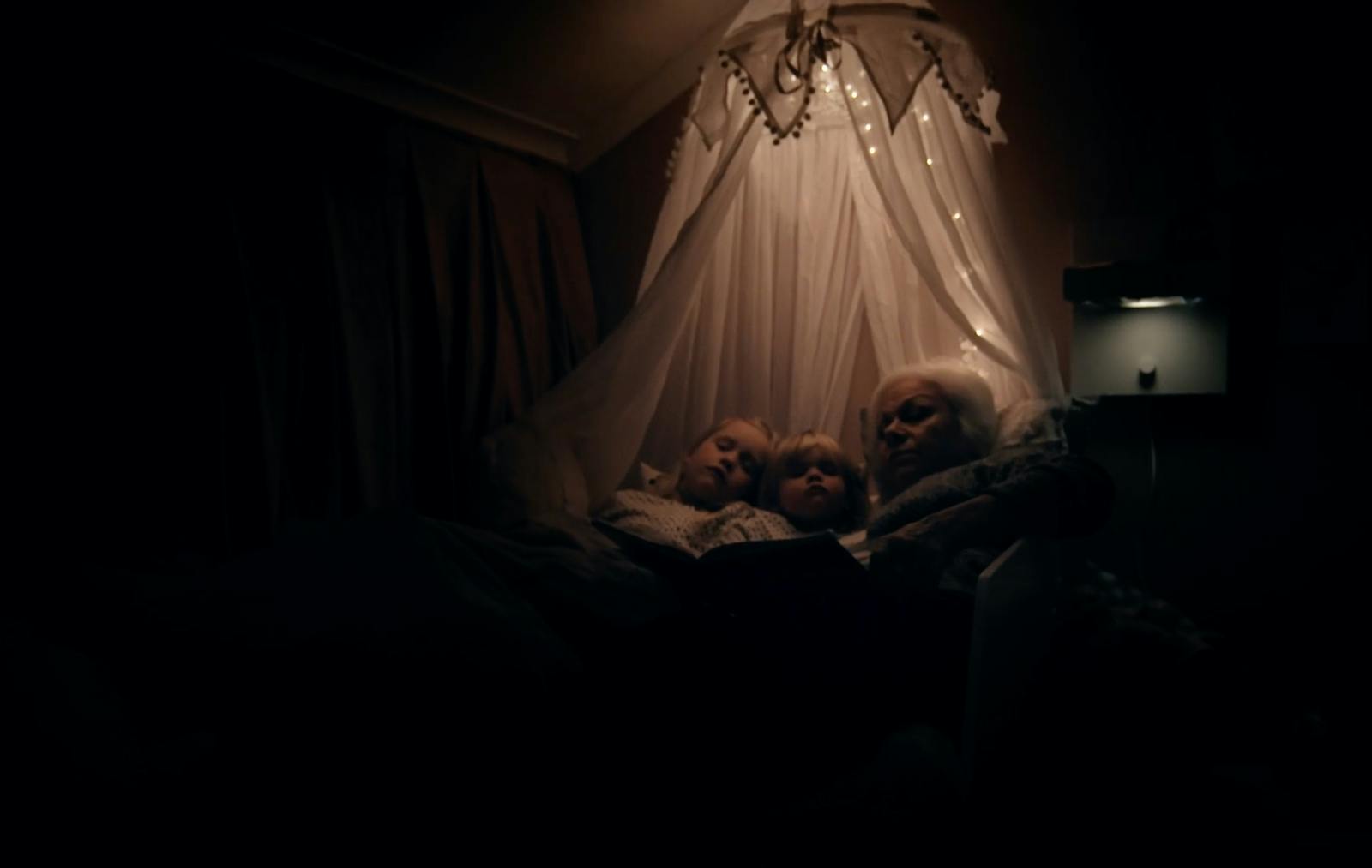 En farmor läser för två barnbarn under en mysigt belyst sänghimmel